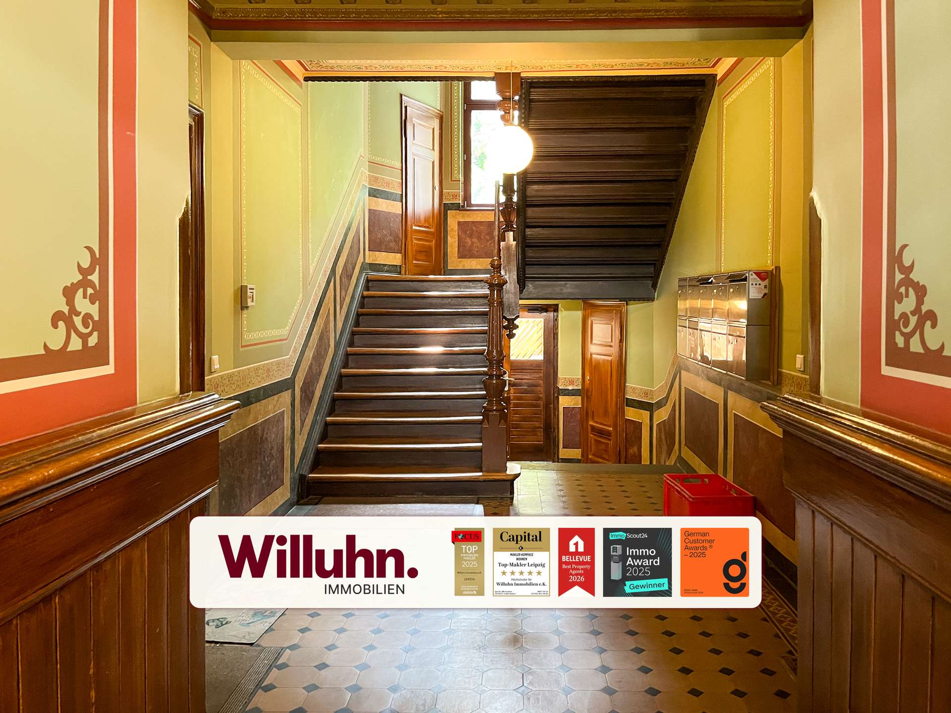 Historic building lobby with a dark wooden staircase, green walls with decorative trim, and a large Willuhn Immobilien banner at the bottom.