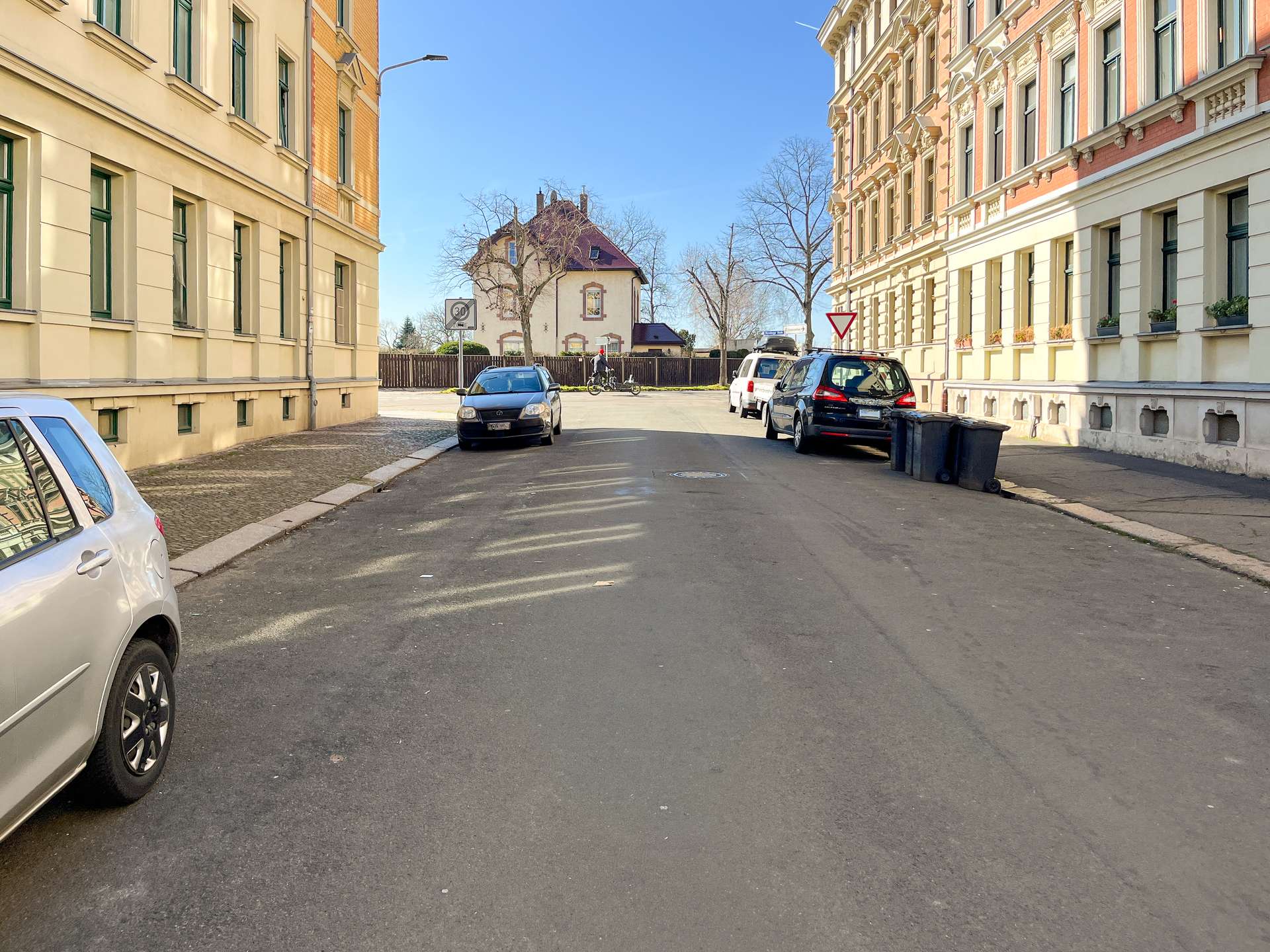 Urban street scene with parked cars along beige and peach buildings, a cyclist in the distance, and a small house at the end under a blue sky.