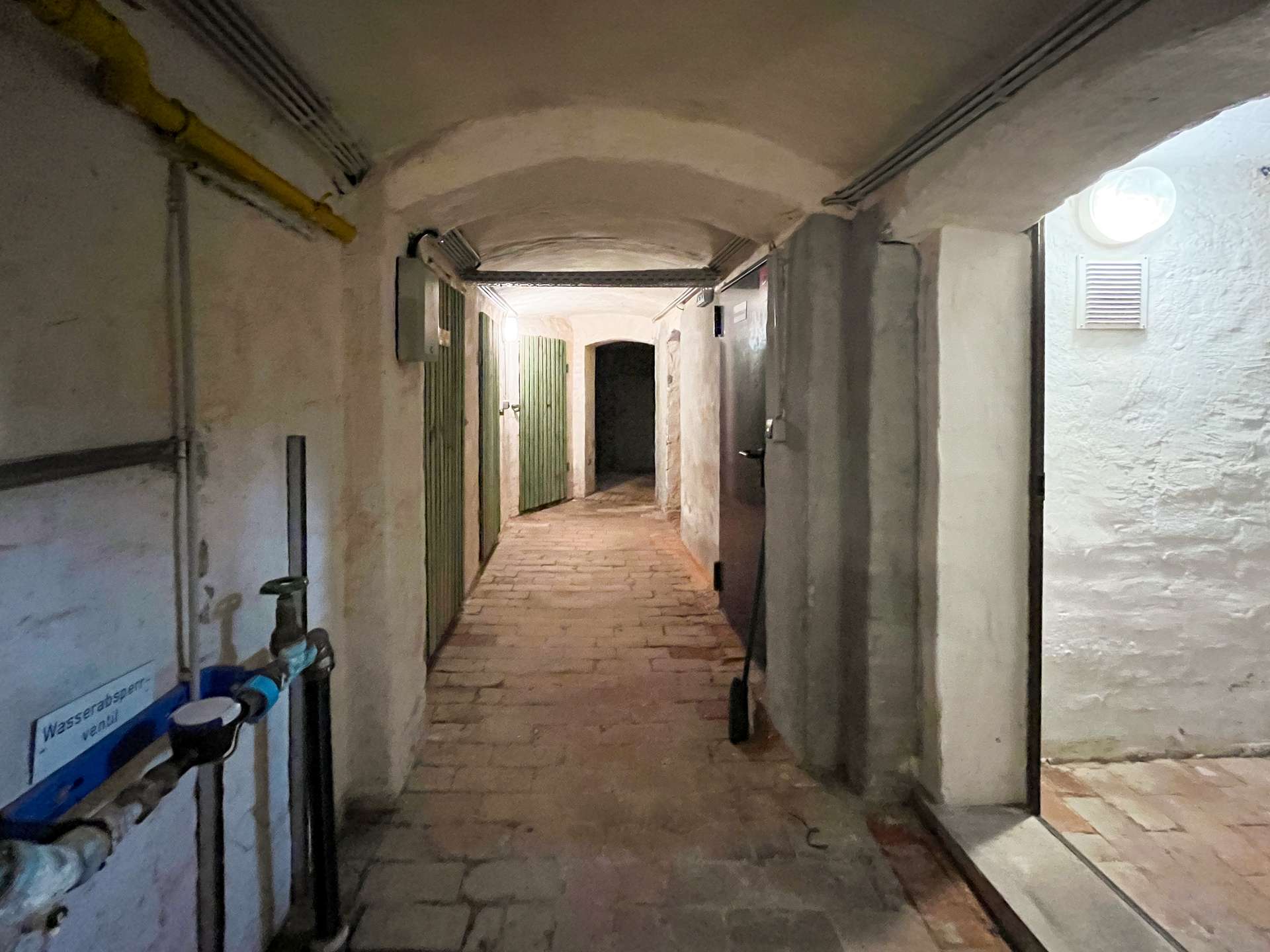 Dimly lit underground corridor with an arched ceiling, stone floor, green metal doors along the left wall, and exposed pipes along the walls; a circular light on the right side wall.
