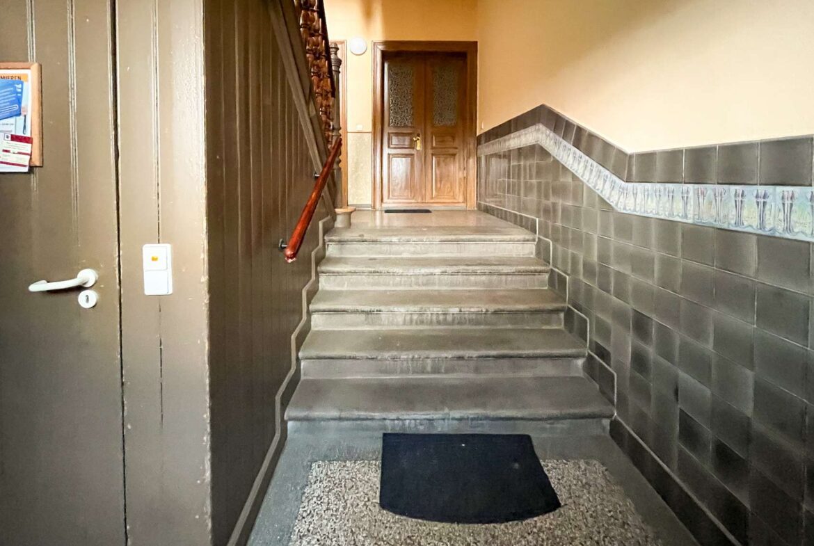 Concrete stairwell with a dark tile wall on the right, leading up to a wooden door at the top of a short flight; a red handrail runs along the left side.