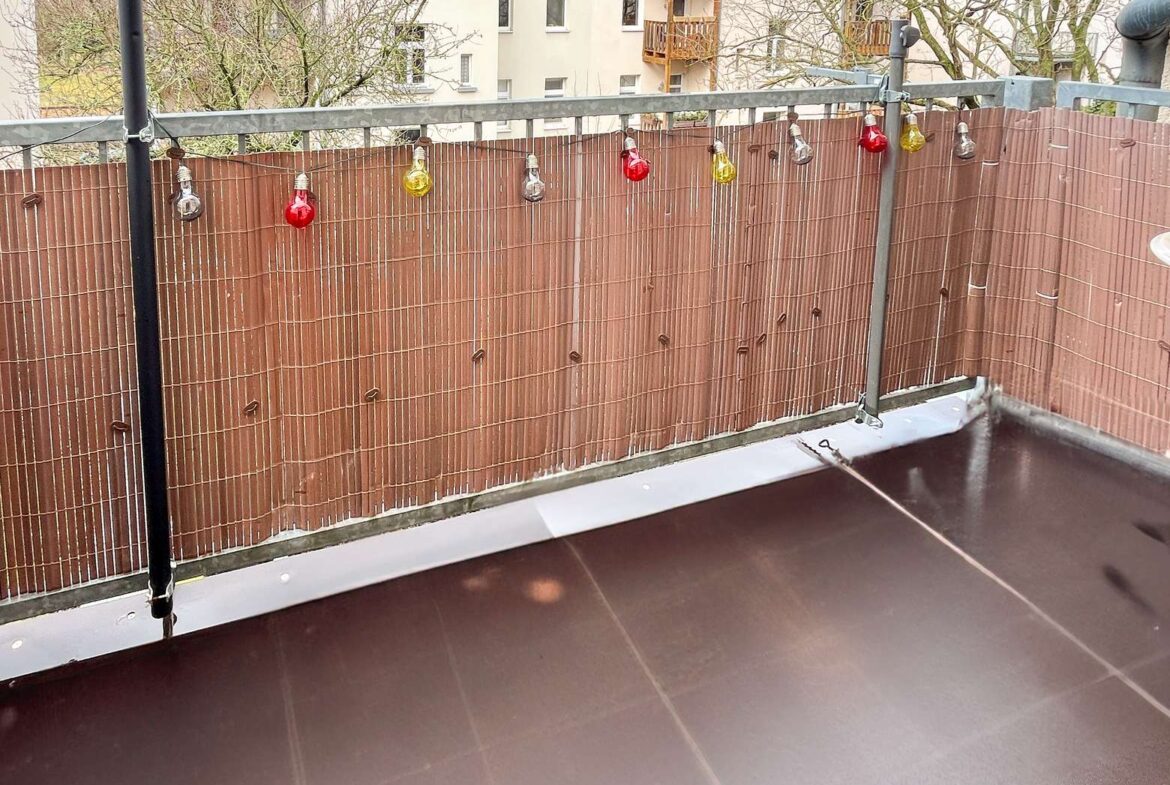 Balcony with a string of colored bulb lights along a brown privacy screen and a wet brown floor; apartment buildings in the background.