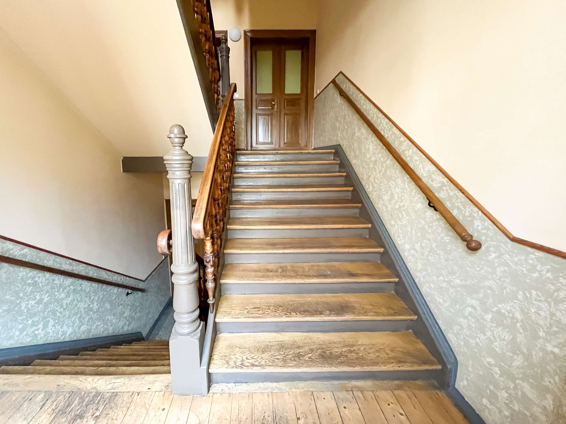 Upward view of a wooden staircase with ornate railing, beige walls, and a closed wooden door at the landing.