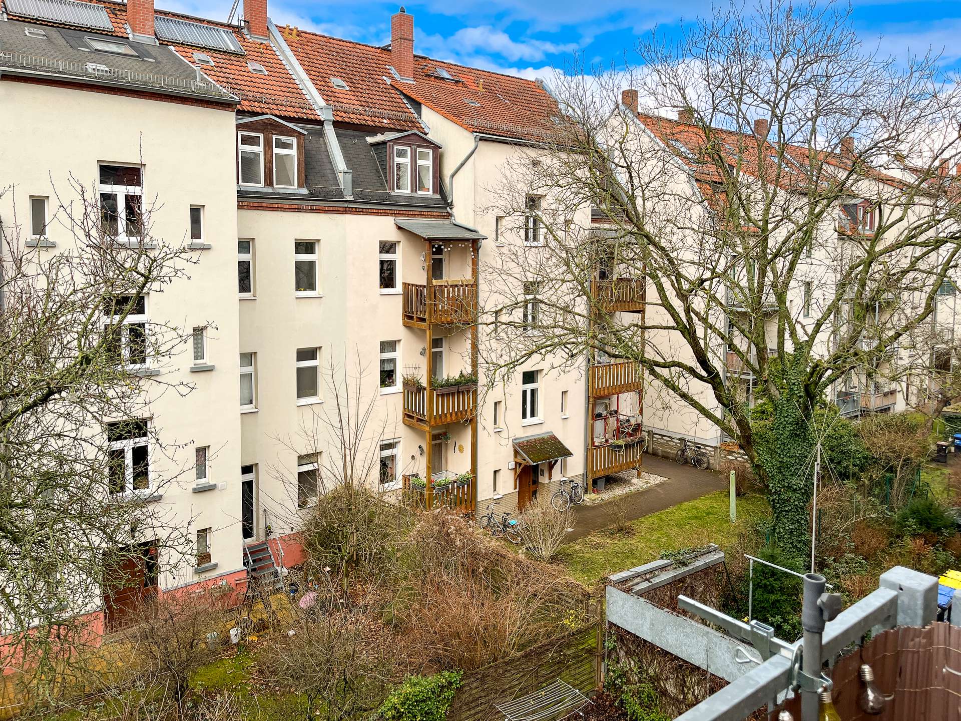 Row of beige apartment buildings with red-tiled roofs, balconies, and leafless trees in a courtyard under a blue sky