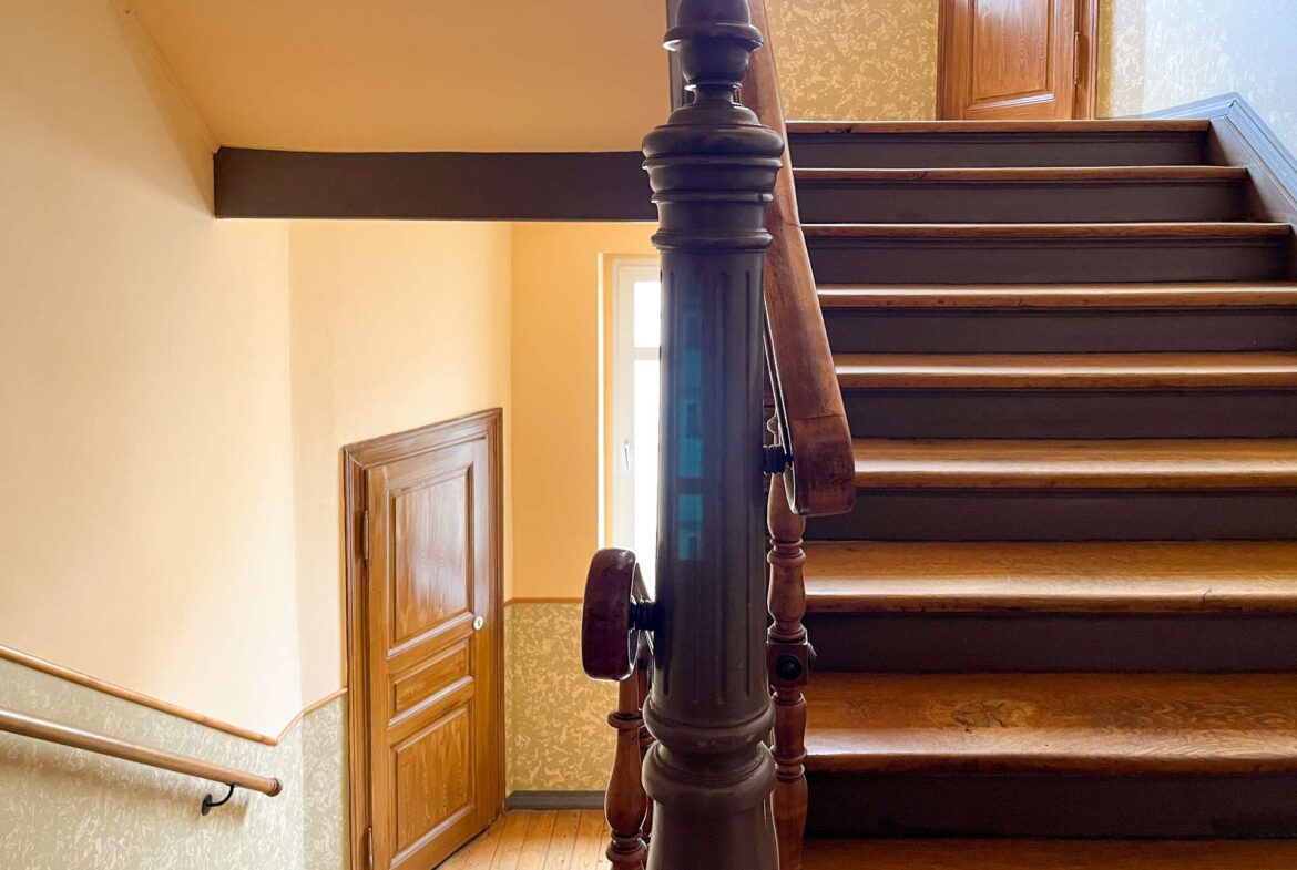 Interior stairwell with a dark wooden banister and ornate newel post, cream walls, and a wooden door at the top.