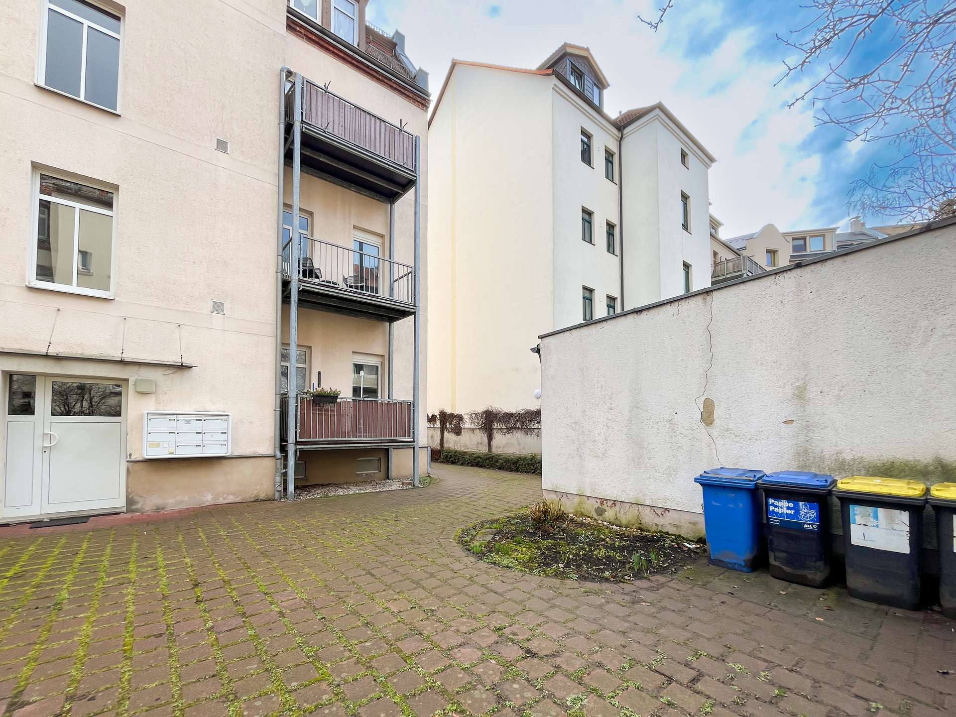 Residential courtyard between beige apartment buildings with balconies, a tall white wall, and blue/black/yellow recycling bins to the right.