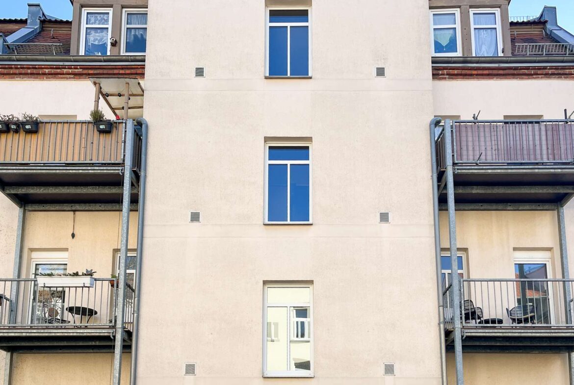 Beige apartment building with a tall central wall, two narrow windows, and balconies on both sides against a blue sky.