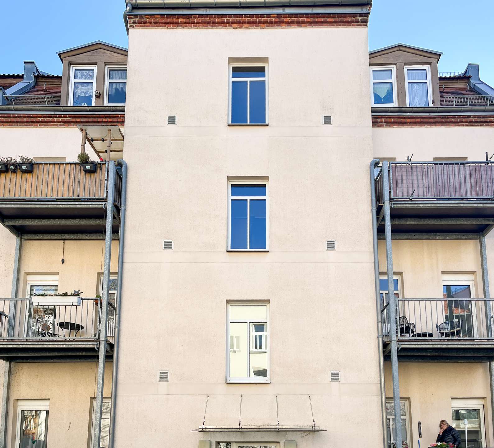 Beige apartment building with a tall central wall, two narrow windows, and balconies on both sides against a blue sky.