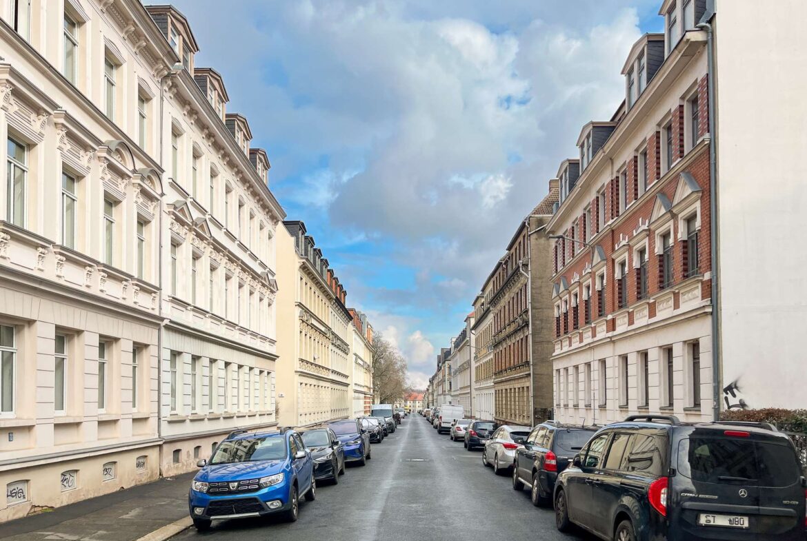 Narrow urban street lined with ornate white and cream townhouses, parked cars on both sides, under a cloudy blue sky.