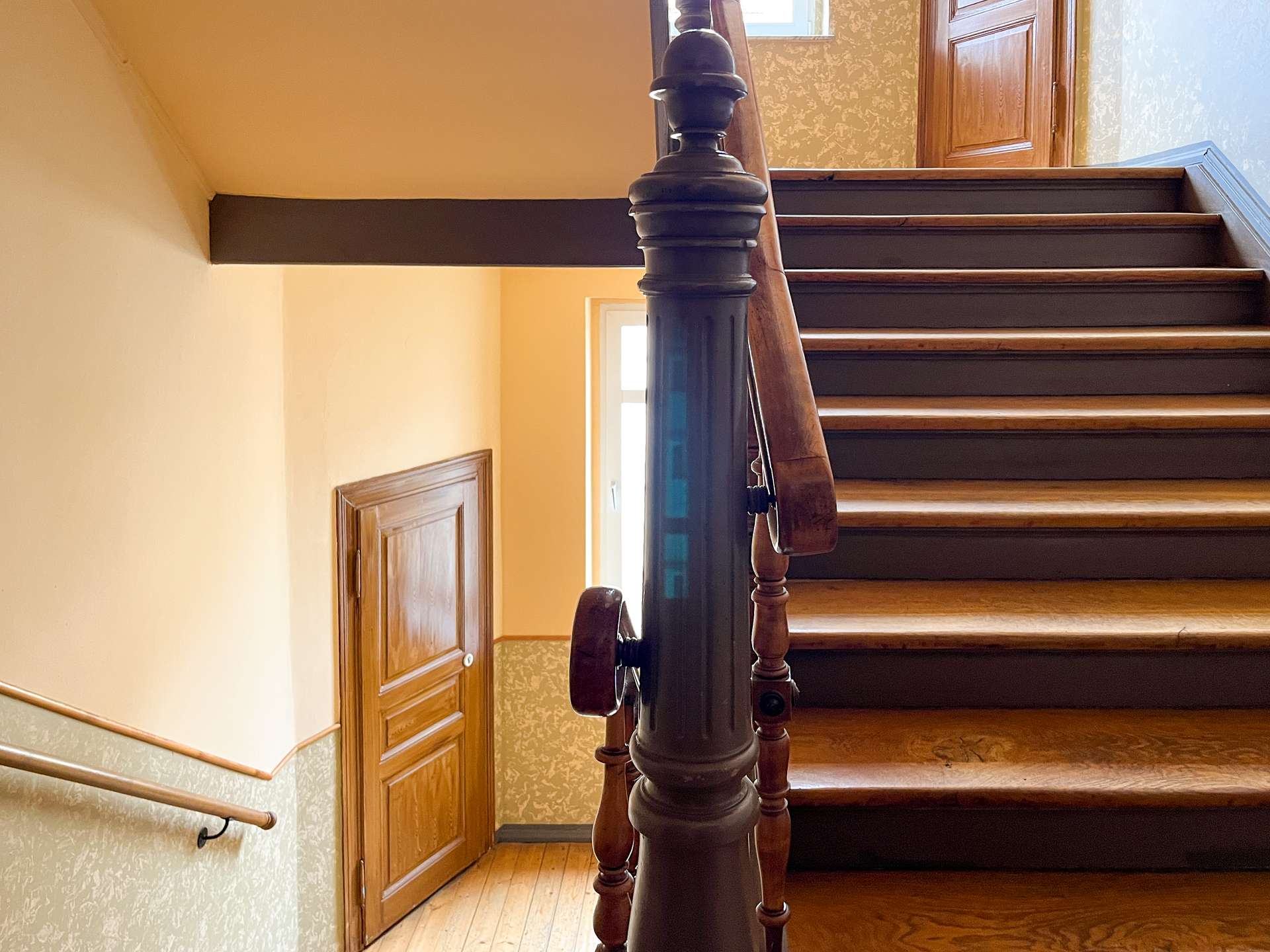 Wooden staircase with a dark green carved newel post and wooden handrail in a warmly lit hallway to the upper floor.