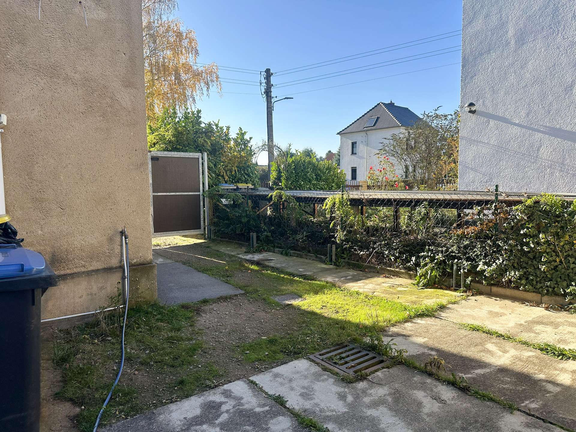 Residential alley with beige building wall on left, chain-link fence, and a white house with a brown roof in the distance under a clear blue sky.