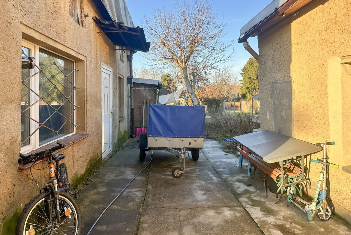 Narrow alley between beige stucco houses with a trailer, bicycles, and miscellaneous outdoor items, and a leafless tree in the background under a blue sky.