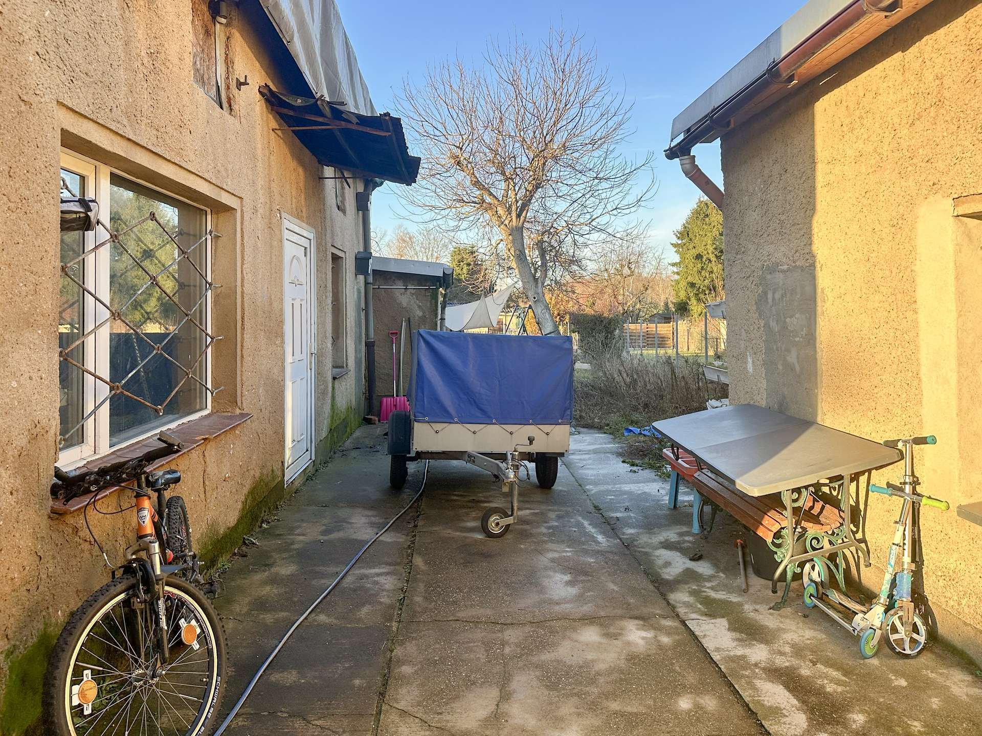 Narrow alley between beige stucco houses with a trailer, bicycles, and miscellaneous outdoor items, and a leafless tree in the background under a blue sky.