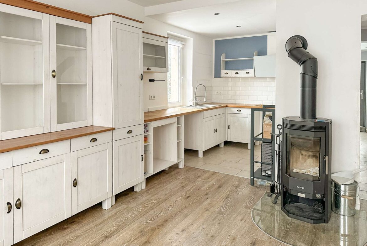 Bright L-shaped kitchen with white cabinets, wooden countertops, and a white subway tile backsplash; a black wood stove stands near curved glass enclosure.
