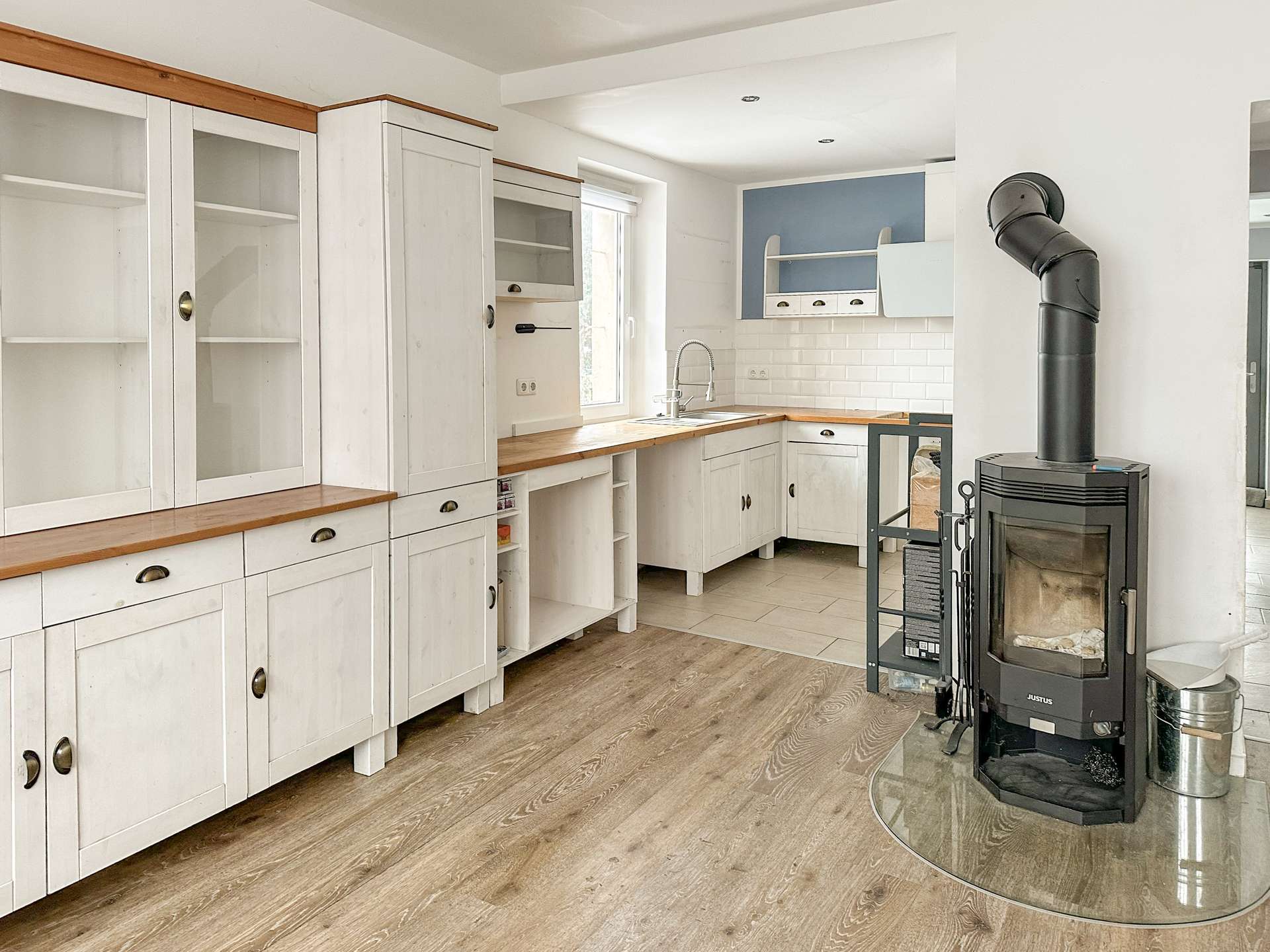Bright L-shaped kitchen with white cabinets, wooden countertops, and a white subway tile backsplash; a black wood stove stands near curved glass enclosure.
