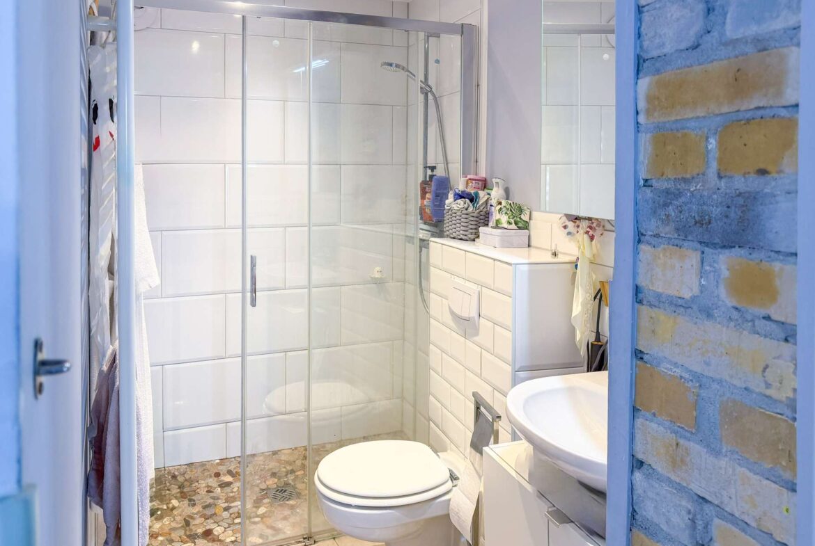 Modern bathroom with glass shower enclosure, white tiled walls, and a toilet and sink; brick accent wall on the right side.