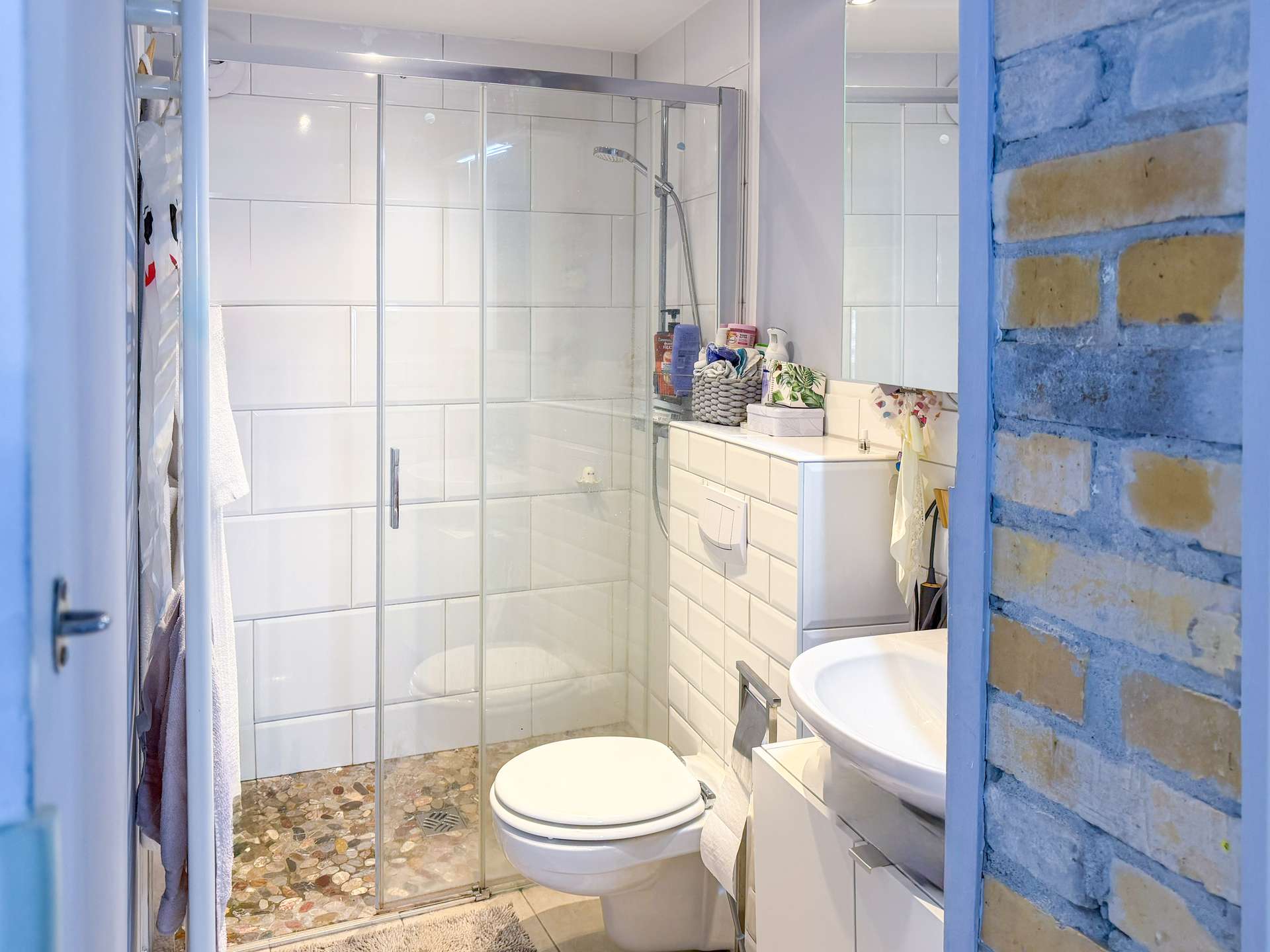 Modern bathroom with glass shower enclosure, white tiled walls, and a toilet and sink; brick accent wall on the right side.