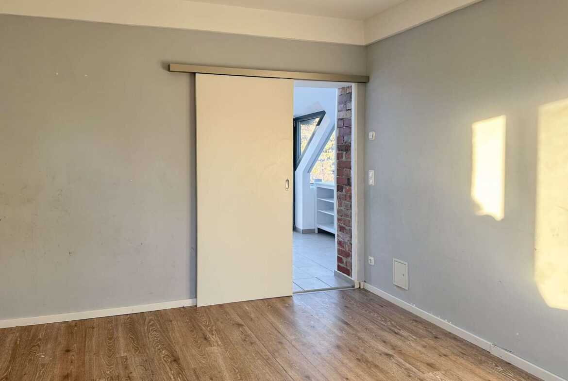 Unfurnished interior room with gray walls, wood-look flooring, and a white sliding door open to a brick-accented opening revealing a stairwell and angled window in the next room.