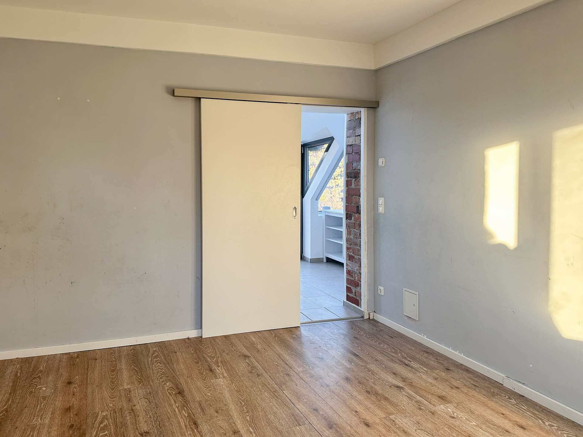 Unfurnished interior room with gray walls, wood-look flooring, and a white sliding door open to a brick-accented opening revealing a stairwell and angled window in the next room.