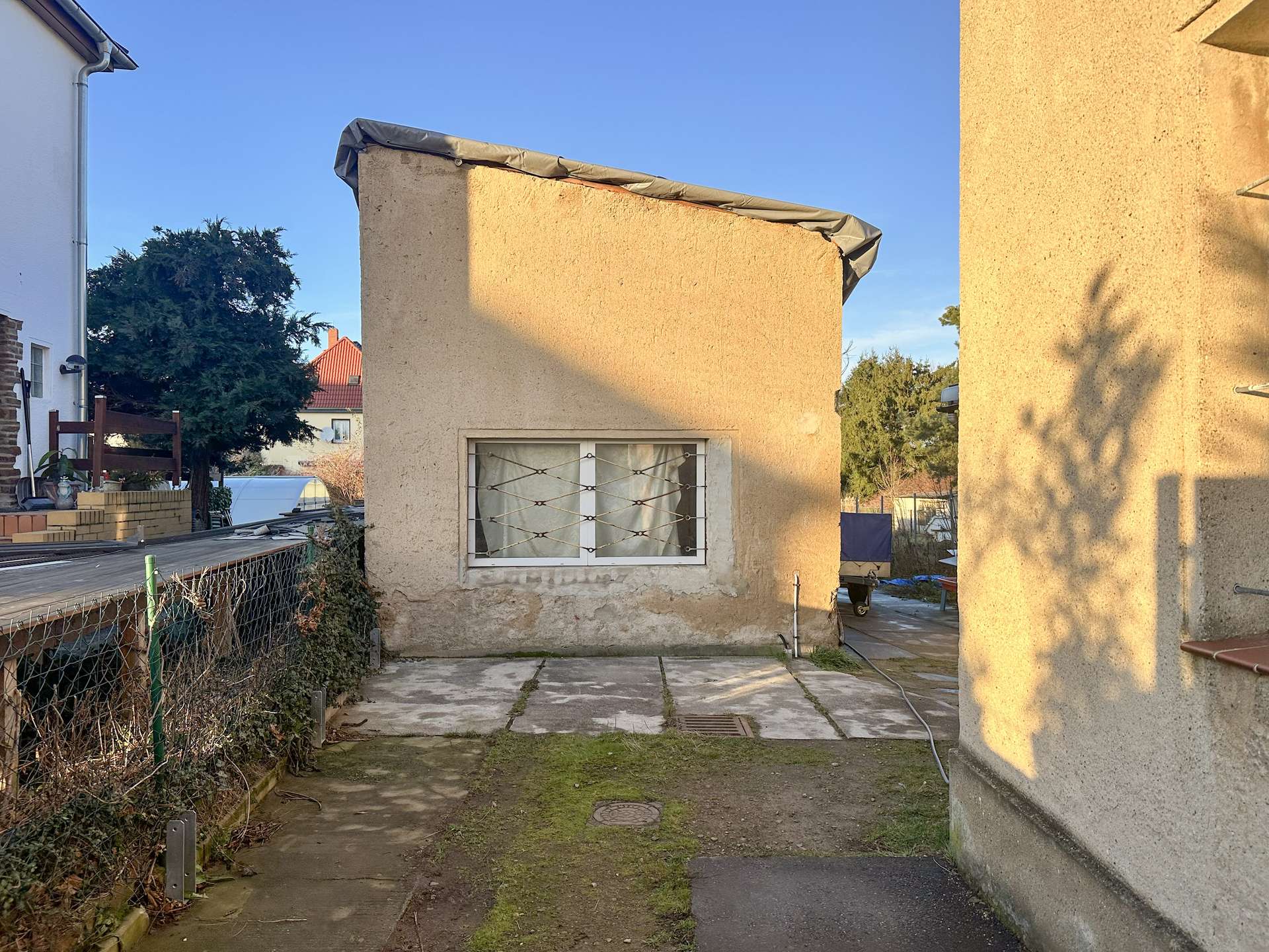 Narrow alley between beige stucco buildings with a small barred window, a tarp along the roof edge, and a chain-link fence on the left.