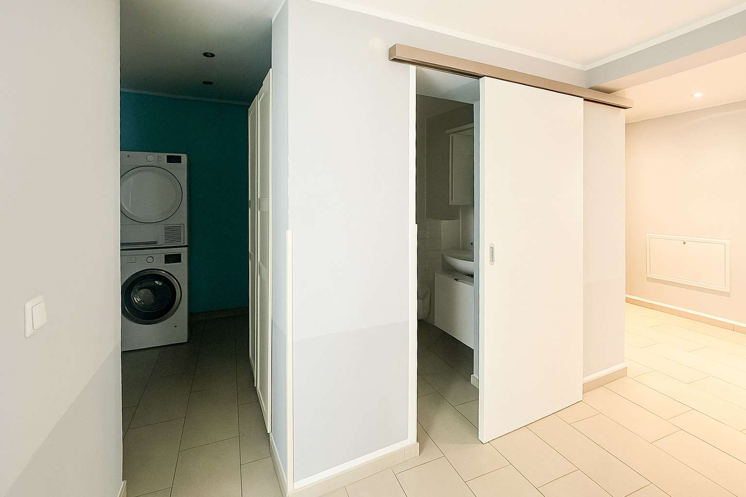 Laundry alcove with stacked washer and dryer against a teal wall, tucked beside white sliding doors in a bright hallway.