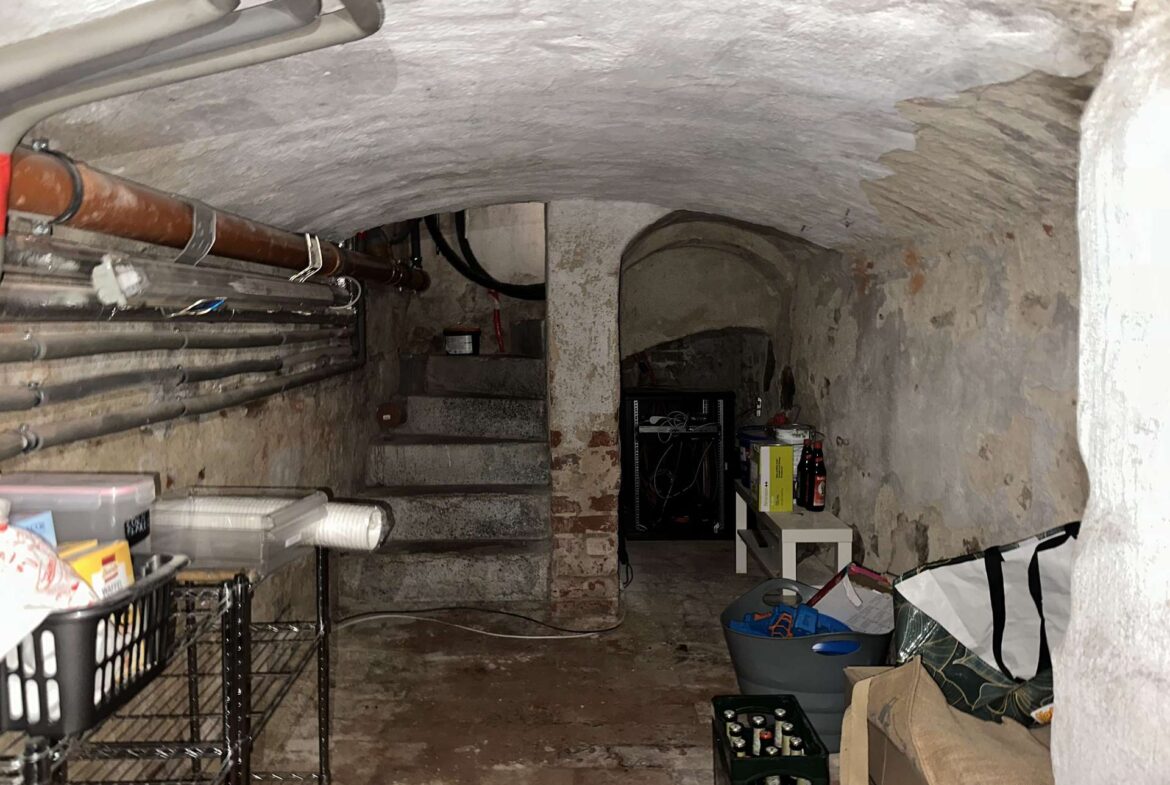 Dimly lit basement storage room with exposed pipes along the left wall, concrete curved ceiling, and cluttered shelves and boxes scattered around.