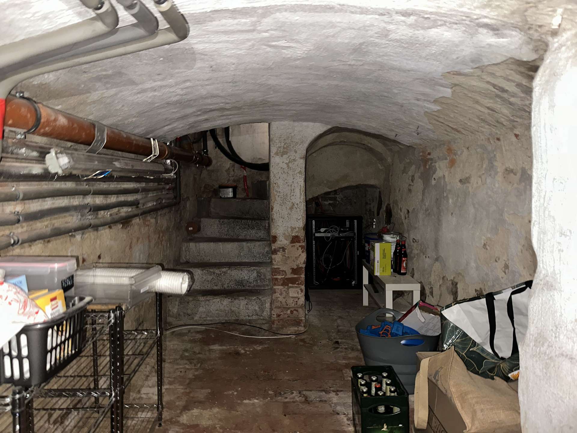 Dimly lit basement storage room with exposed pipes along the left wall, concrete curved ceiling, and cluttered shelves and boxes scattered around.