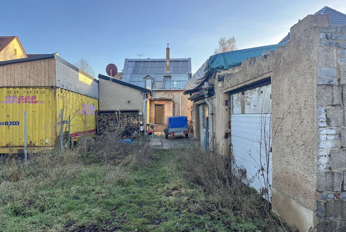 Narrow alley between rundown buildings, yellow DHL container wall on the left, stacked firewood and a small trailer in the center, with a partially ruined right wall and blue sky above.