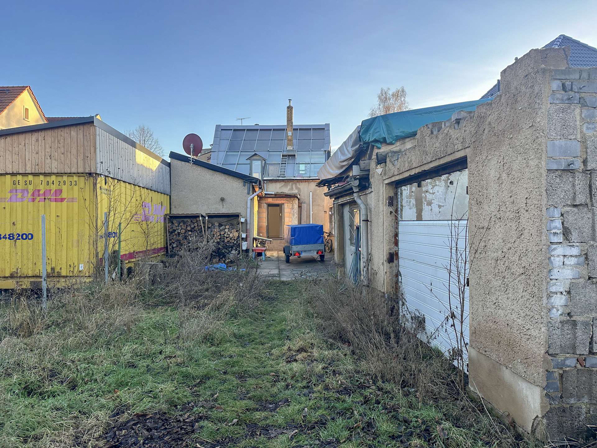 Narrow alley between rundown buildings, yellow DHL container wall on the left, stacked firewood and a small trailer in the center, with a partially ruined right wall and blue sky above.