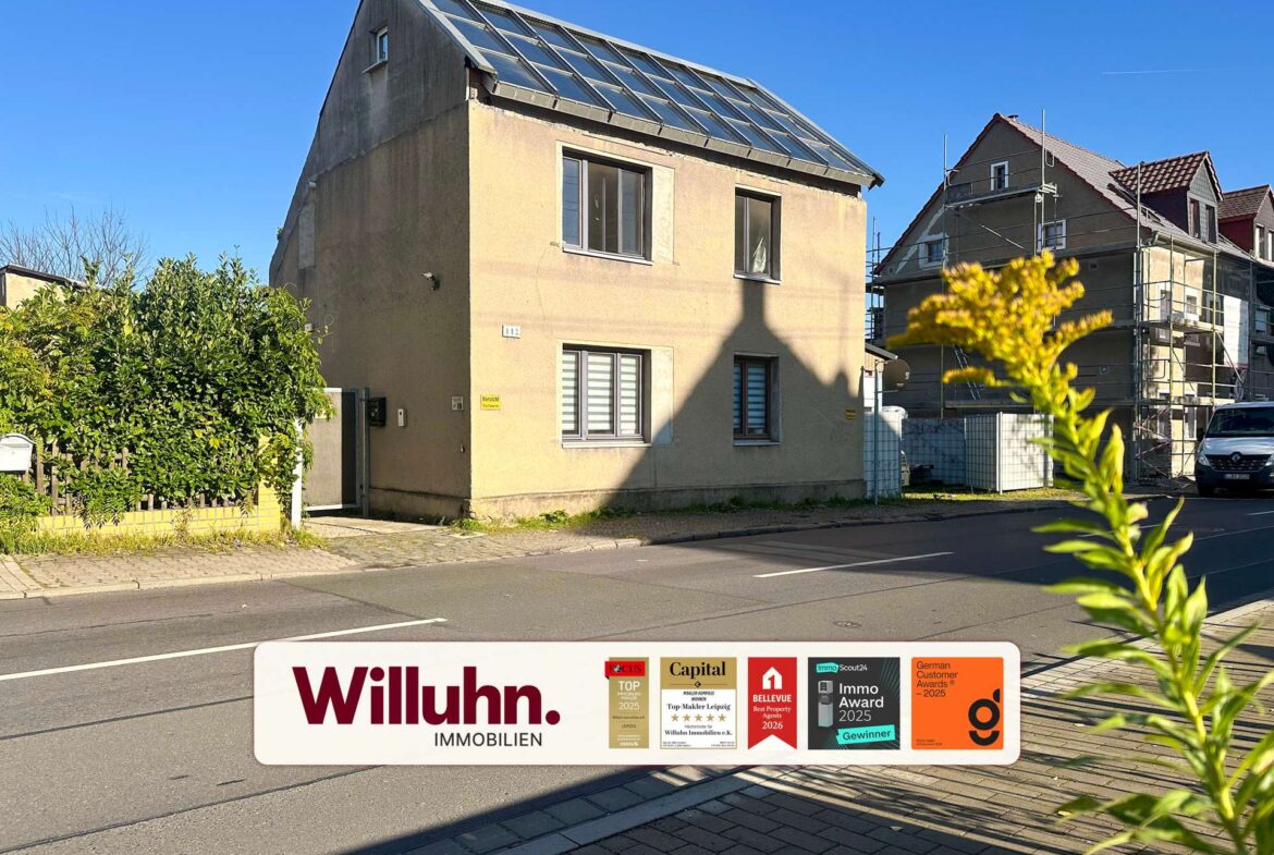 Front view of a beige house on a sunny street with a large real estate sign in the foreground showing 'Willuhn Immobilien' and awards stickers.