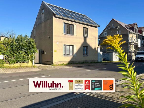 Front view of a beige house on a sunny street with a large real estate sign in the foreground showing 'Willuhn Immobilien' and awards stickers.
