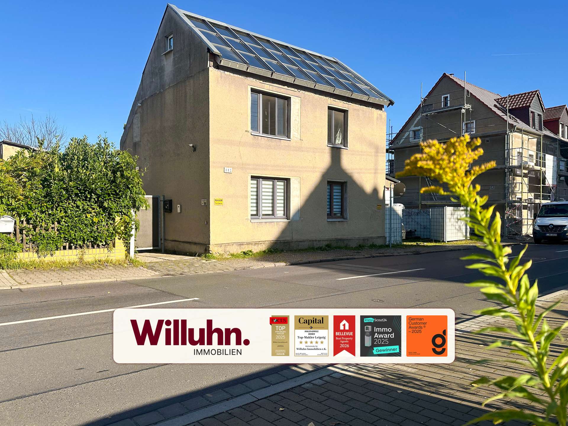 Front view of a beige house on a sunny street with a large real estate sign in the foreground showing 'Willuhn Immobilien' and awards stickers.