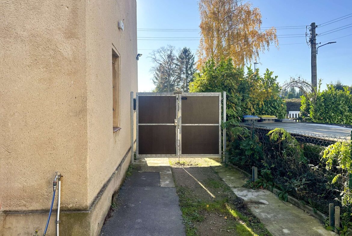 Narrow outdoor alley beside a beige stucco building with a metal gate and overgrown greenery on the right; blue sky above.