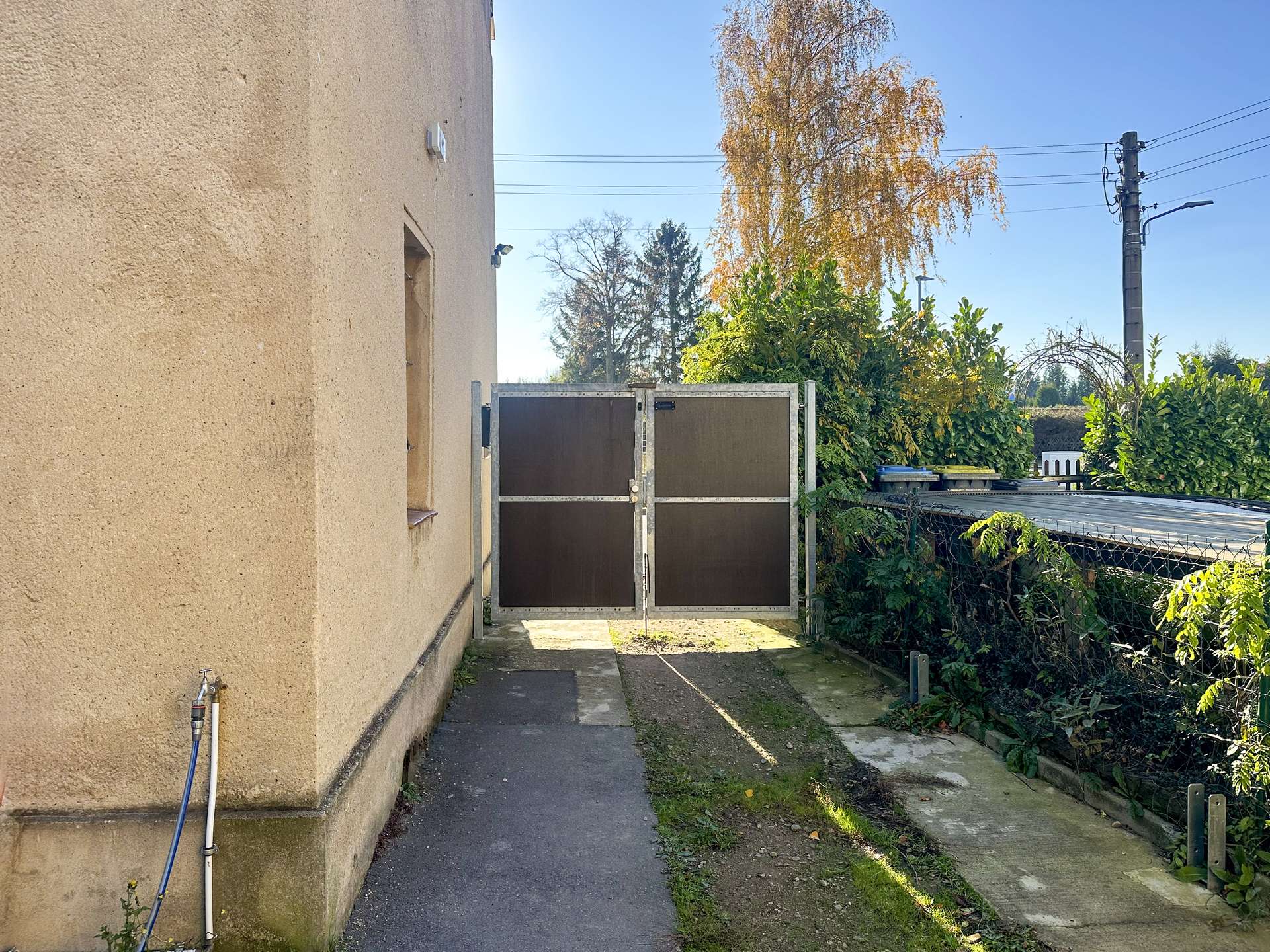 Narrow outdoor alley beside a beige stucco building with a metal gate and overgrown greenery on the right; blue sky above.