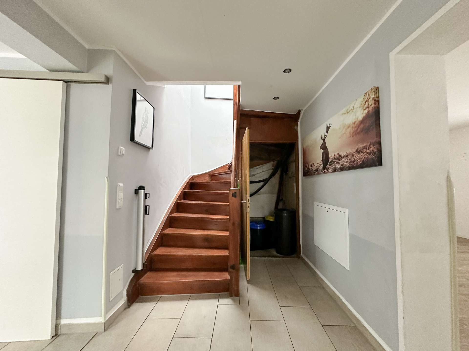 Interior hallway with a wooden staircase on the left, gray walls, and a large deer photo on the right wall; storage under the stairs.