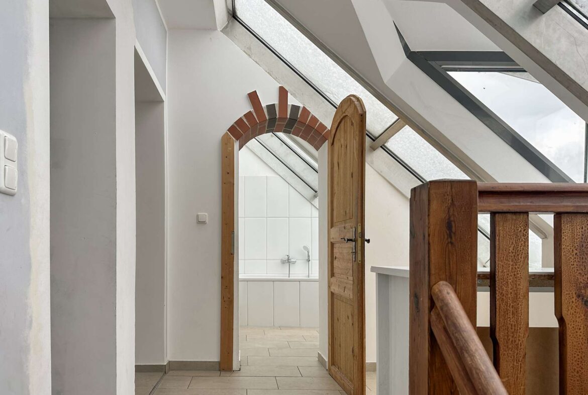 Open wooden door leads to a bright bathroom with white tiles, seen from a hallway with skylight windows above (attic space).