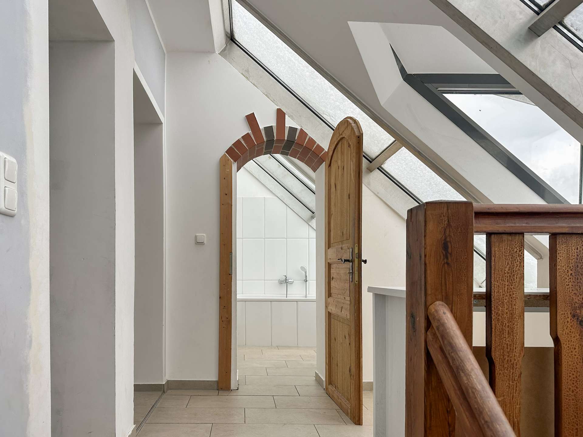 Open wooden door leads to a bright bathroom with white tiles, seen from a hallway with skylight windows above (attic space).