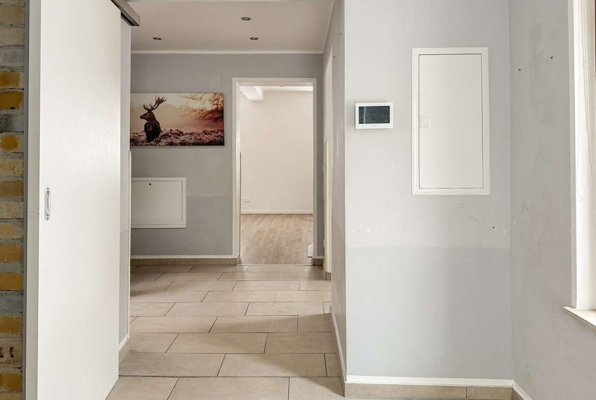 Hallway with gray walls and beige tile floor, open door to a room with a deer painting; white cabinet on the left and a wall control panel on the right.