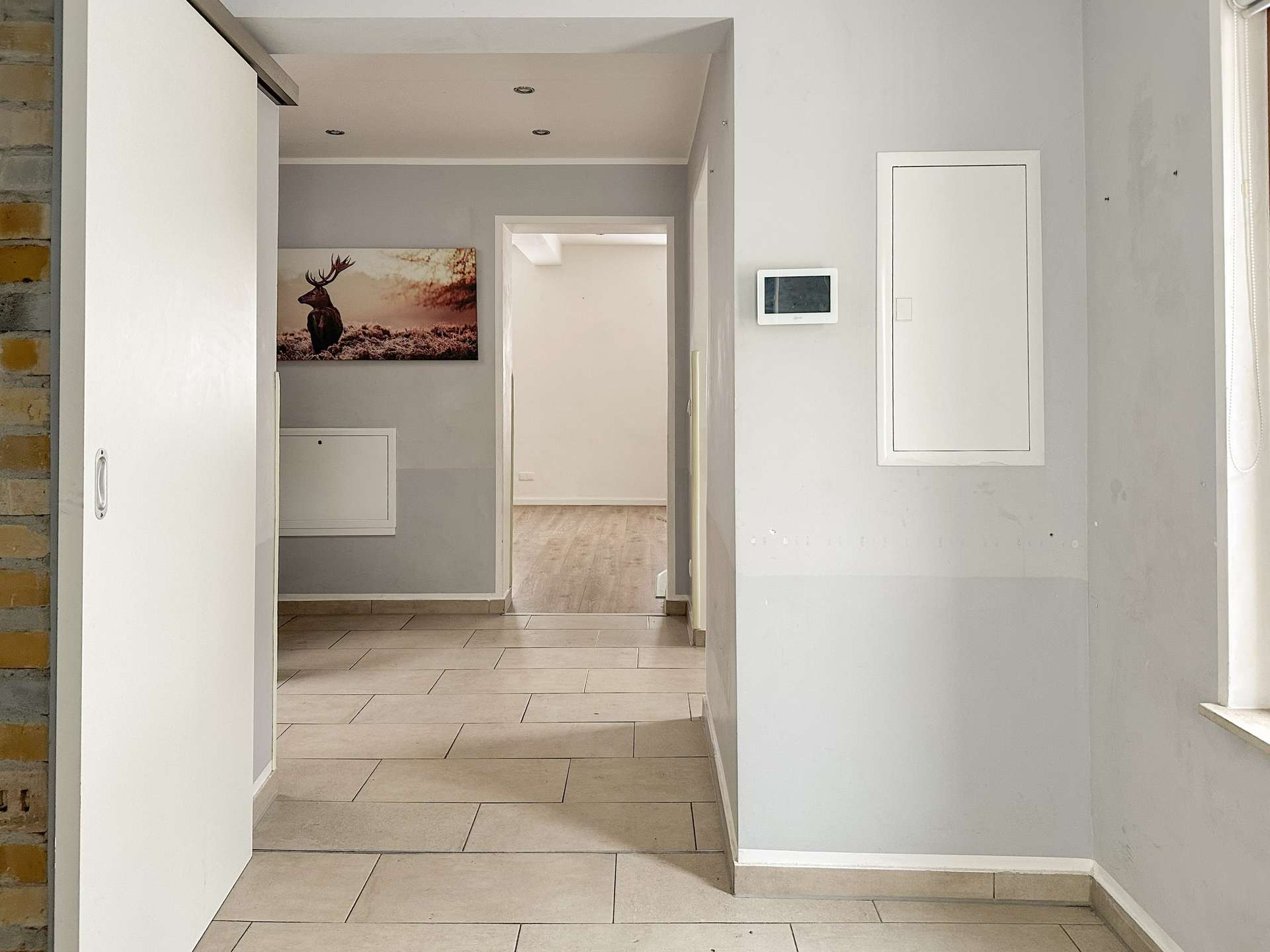 Hallway with gray walls and beige tile floor, open door to a room with a deer painting; white cabinet on the left and a wall control panel on the right.
