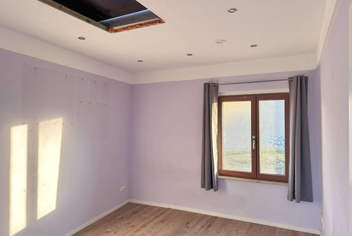 A small purple room with a window, gray curtains, and sunlight casting a rectangular glow on the wall; ceiling has a large open hole.