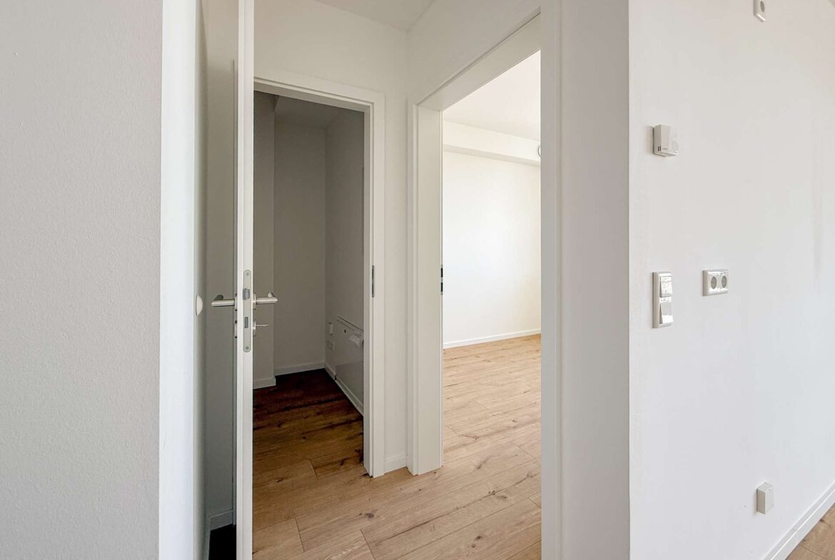 Interior hallway with white walls and light wood flooring; an open door reveals a bright, empty room.