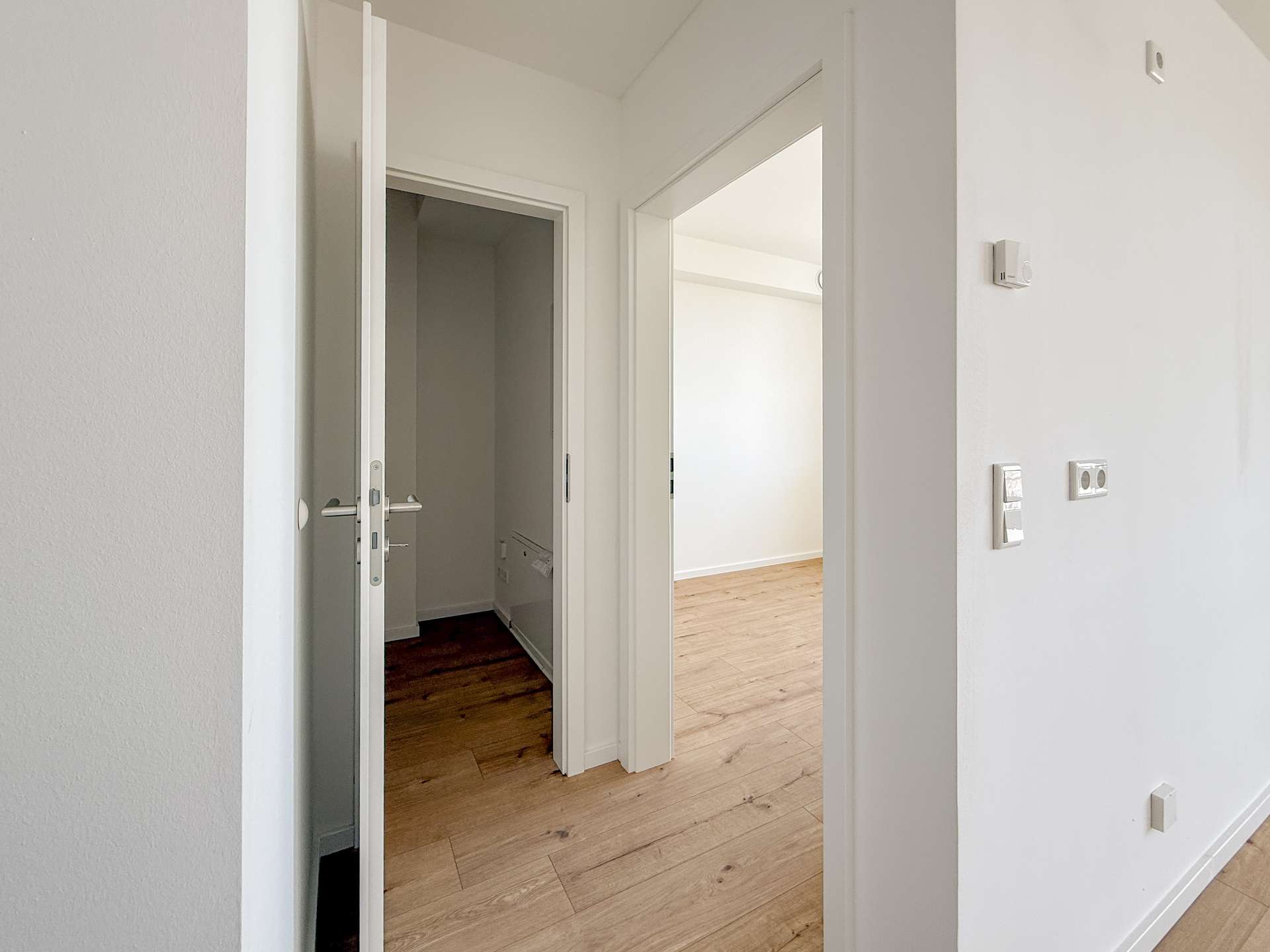 Interior hallway with white walls and light wood flooring; an open door reveals a bright, empty room.