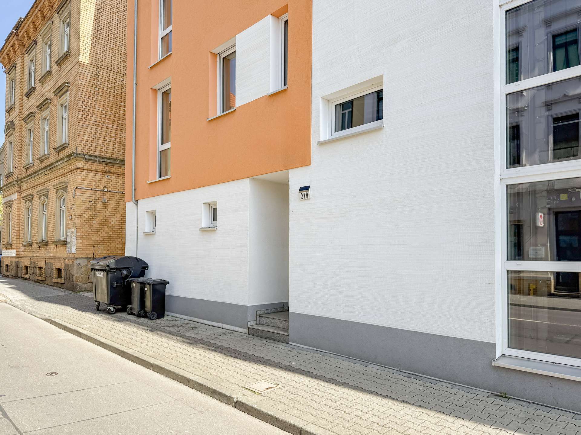 Street view of a modern orange-and-white residential building beside an older brick building; trash bins on the sidewalk in front.