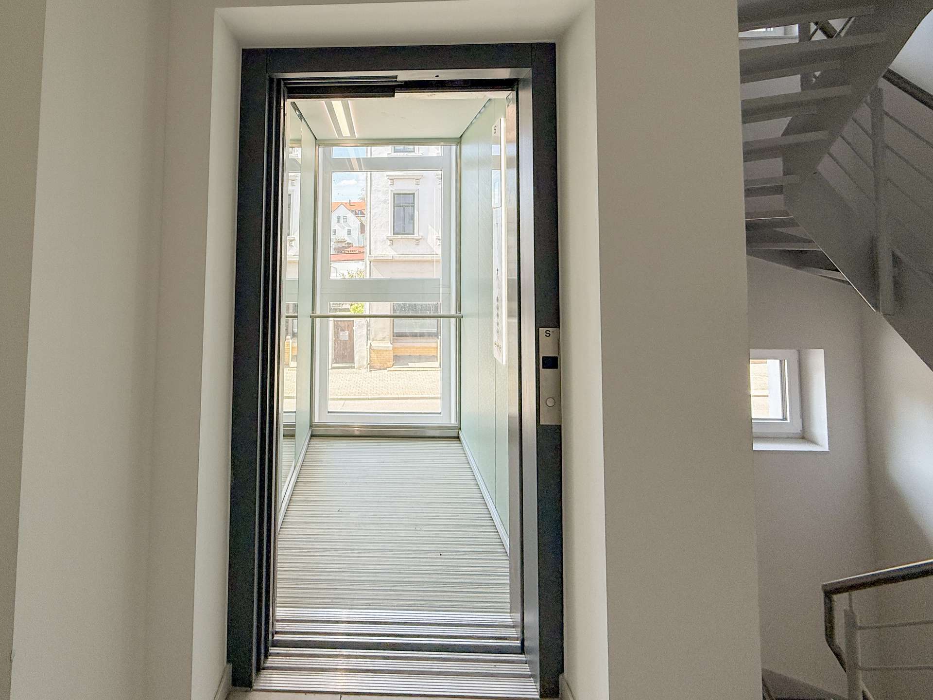 Open elevator doors show a narrow glass shaft with a street view of buildings outside, hallway walls on either side, and a metal floor grate at the threshold.
