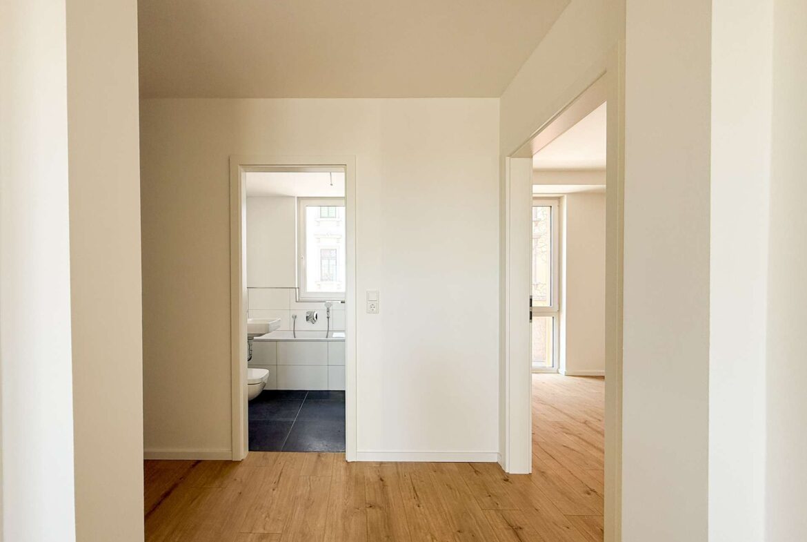 Bright hallway with pale walls and warm wood flooring; a doorway on the left opens to a bathroom with white tiles and a window visible beyond.
