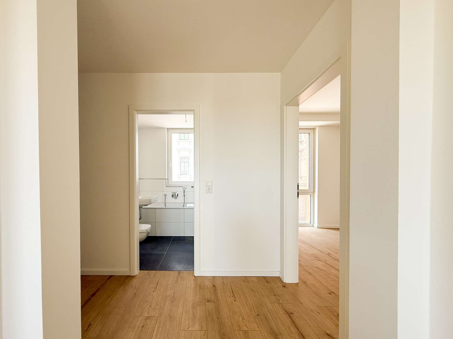 Bright hallway with pale walls and warm wood flooring; a doorway on the left opens to a bathroom with white tiles and a window visible beyond.