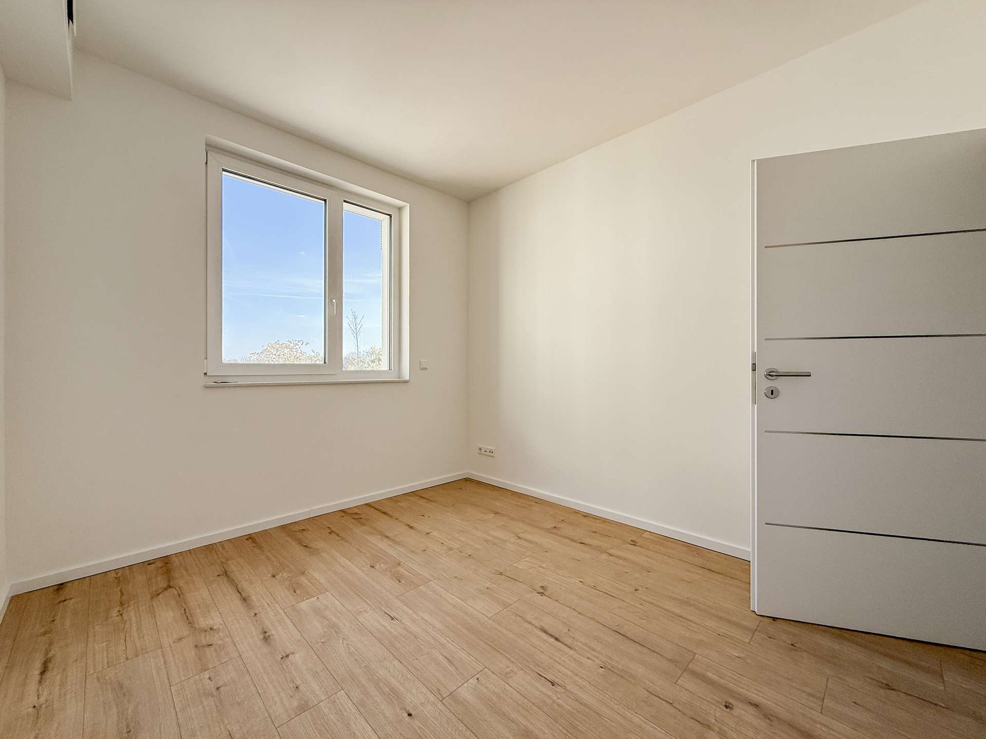 Empty white room with light wood floor, a window letting in daylight, and a grey door with horizontal grooves on the right.