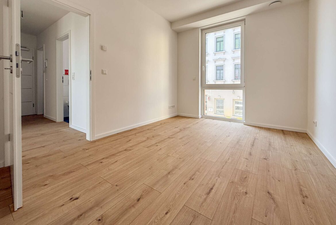 Empty sunlit room with light wood flooring and a large window overlooking a city street outside the window area on the right side of the image.