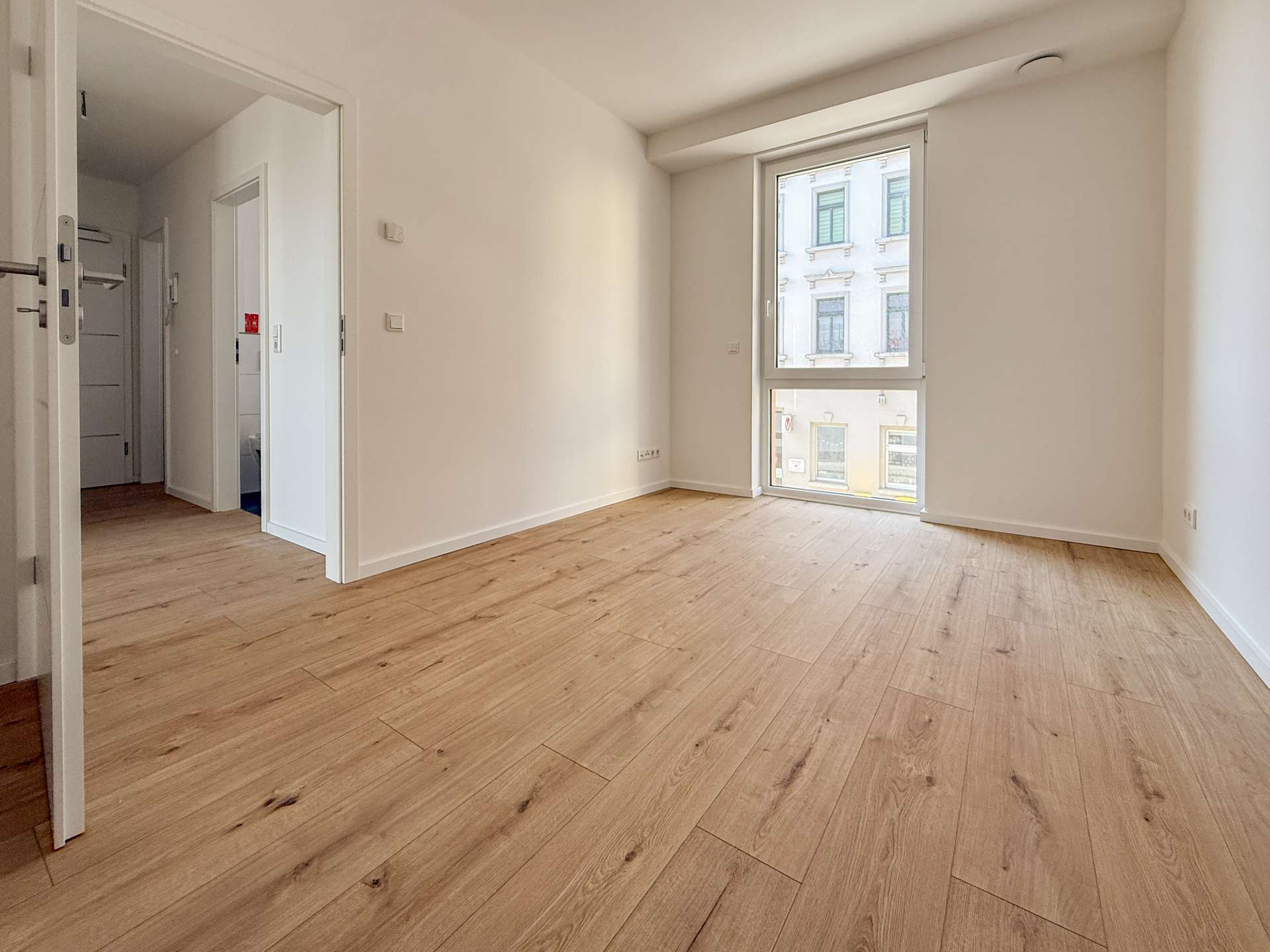 Empty sunlit room with light wood flooring and a large window overlooking a city street outside the window area on the right side of the image.
