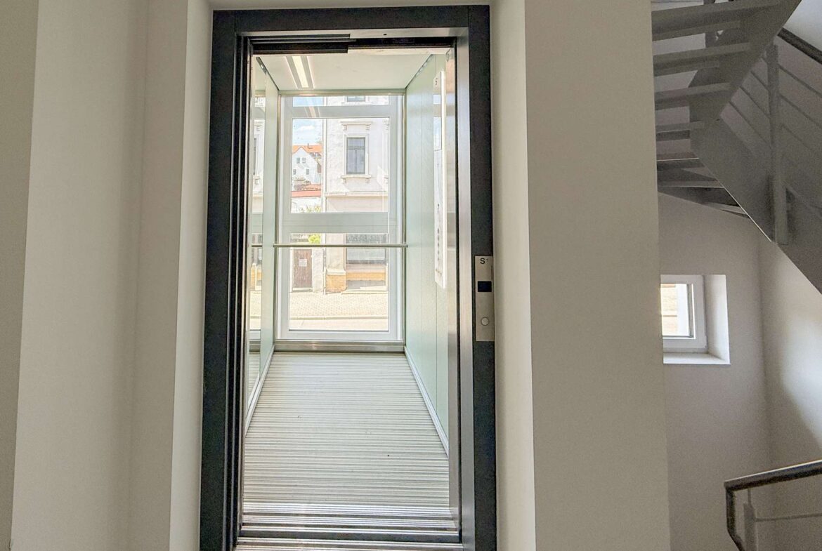 Open elevator doors with a glass enclosure; view of urban buildings outside through the glass panels at the far end of the hall.