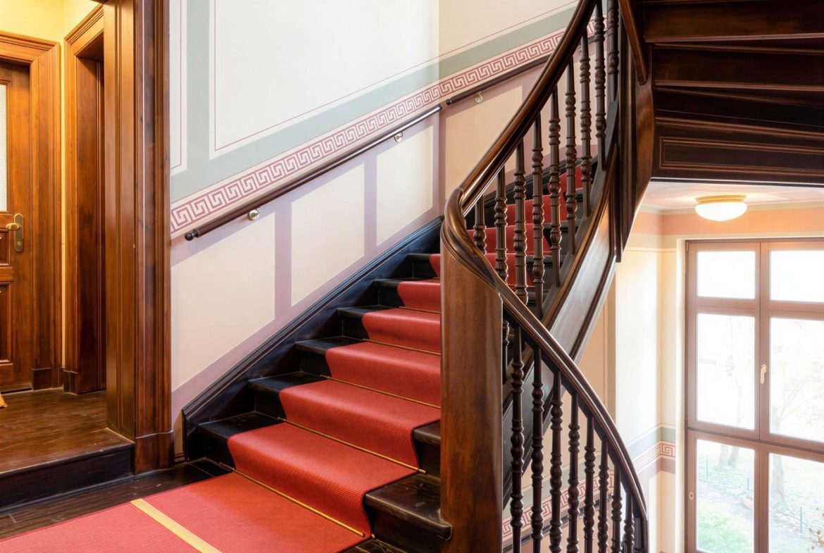 Curved wooden staircase with a red carpet, ornate balusters, and panelled walls in a sunlit foyer.